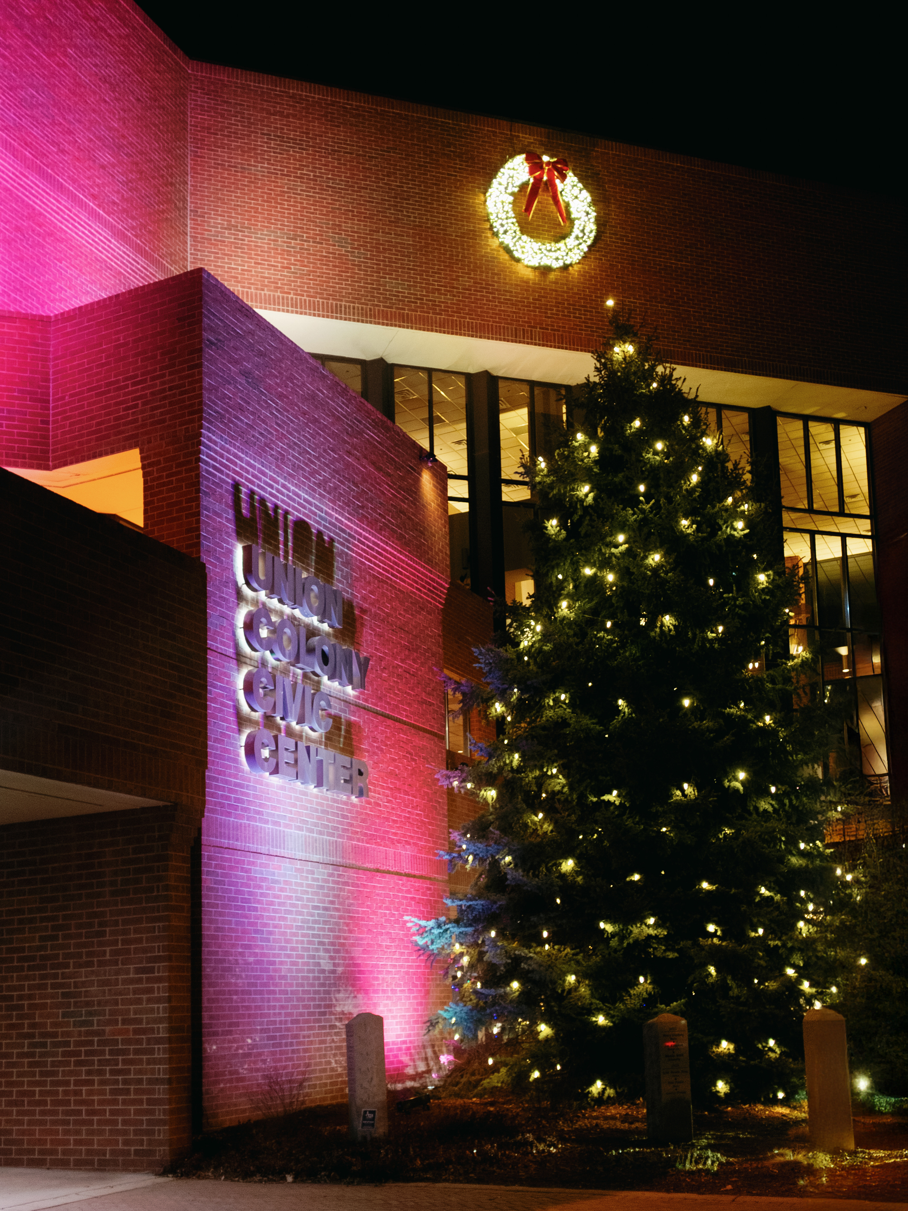 Outside building of UCCC, signage and brick lit with festive red and purple, while a white light wreath with red bow sits higher on the building. The building is lit from the interior, and a large evergreen is adorned with white Christmas lights.