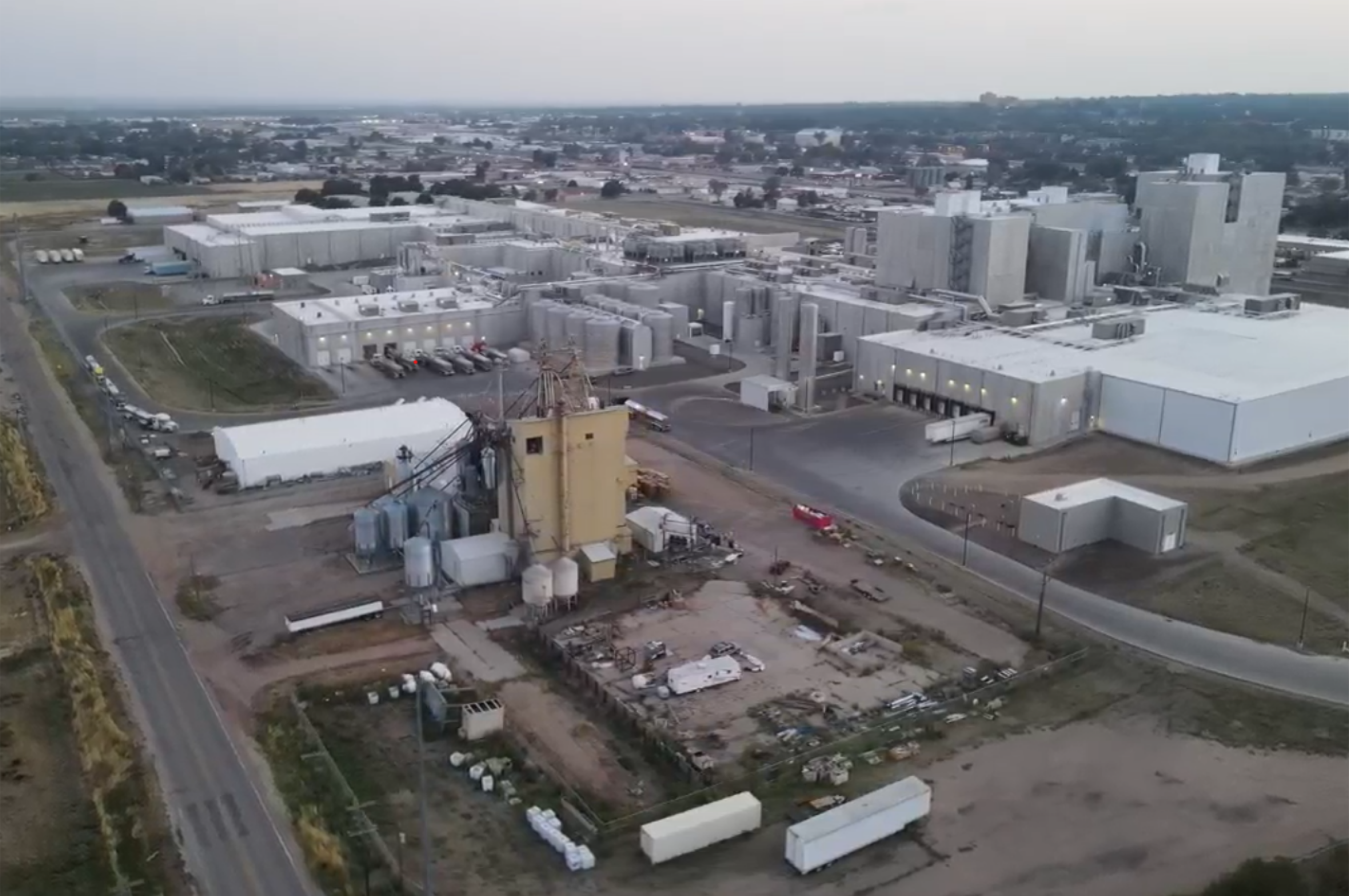This image depicts an aerial view of Leprino Foods, one of Greeley's primary employers.  The image shows a large industrial facility with multiple warehouse-style buildings and storage tanks, surrounded by roads and open land. The complex includes a central plant area with pipes and silos, and several trailers and vehicles are visible on the property.  
