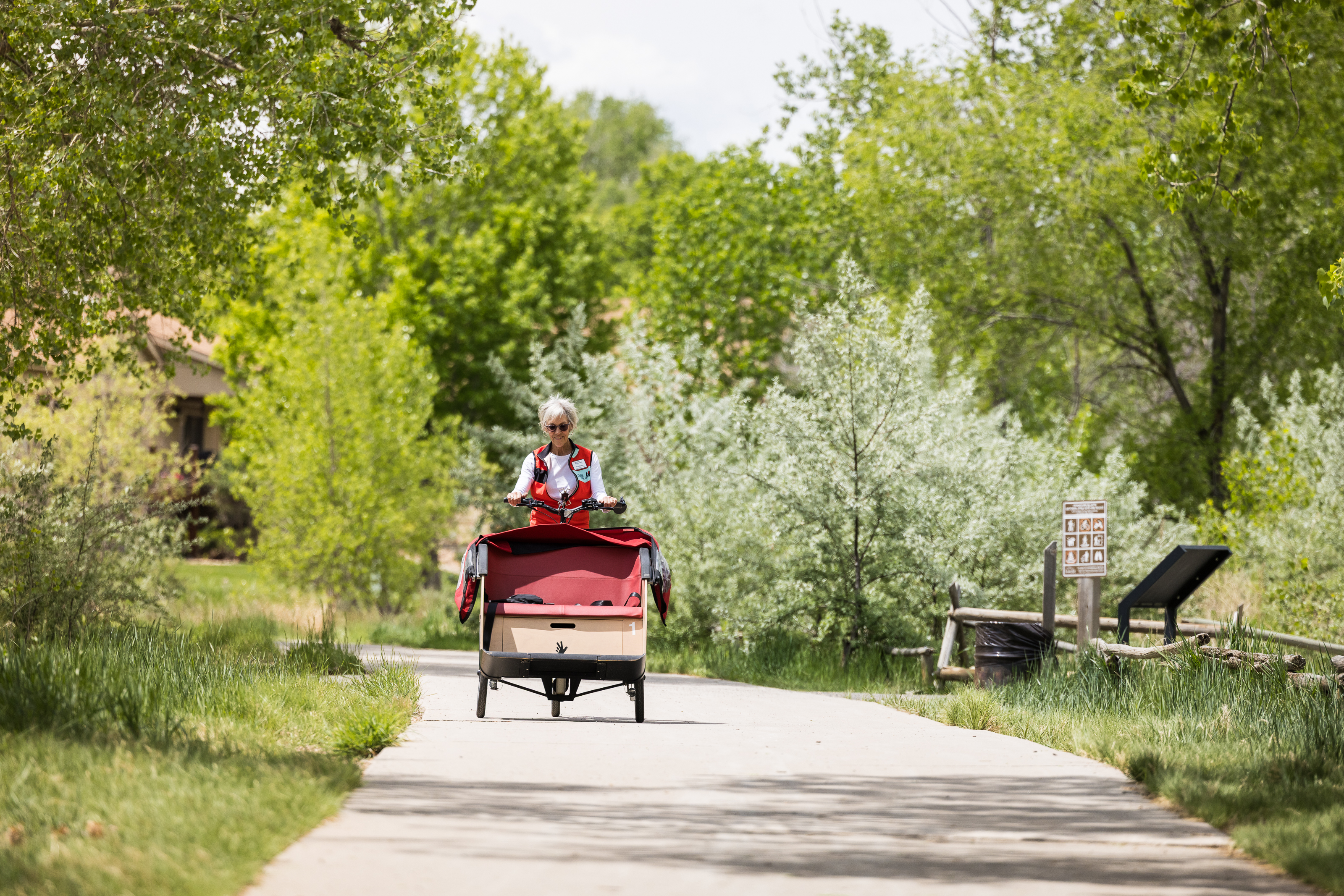 Cyclist riding a red three-wheeled bicycle on a paved trail surrounded by lush green trees and shrubs in Greeley.