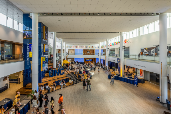 Wide view of the University of Northern Colorado Campus Commons atrium with students gathered near stairs, seating areas, flags, and campus displays.