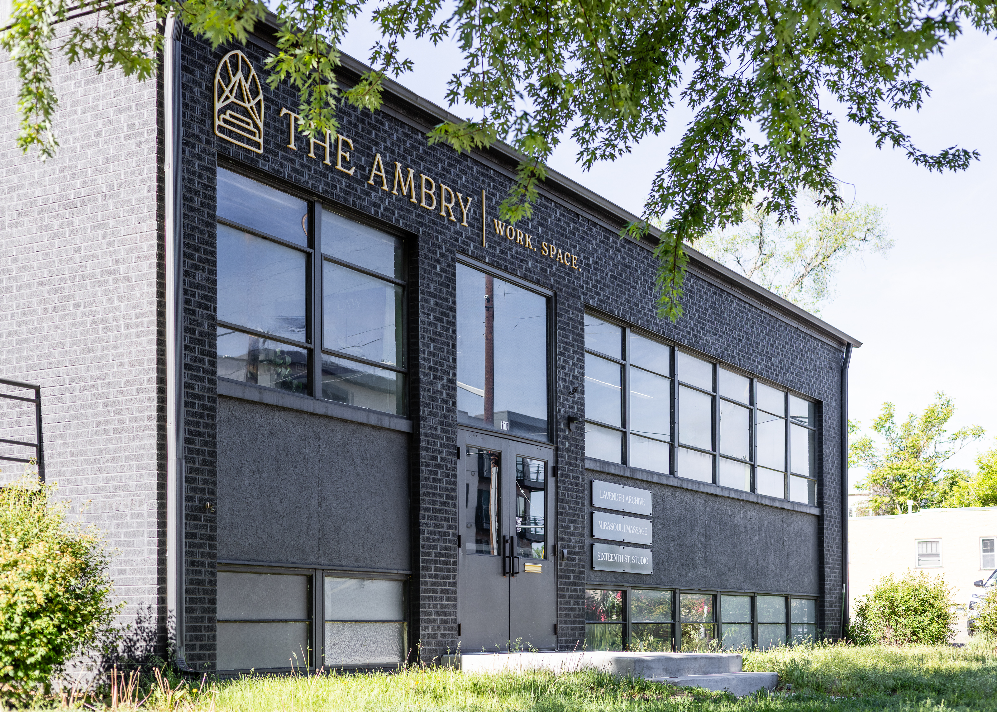This image depicts The Ambry coworking space used by Greeley startup businesses and entrepreneurs.  The building is a rectangular, two-story facility with a black brick facade and ample glass windows on both floors.  The front entrance double-doors have large glass windows above, and the building is surrounded by grass, trees, and green bushes.