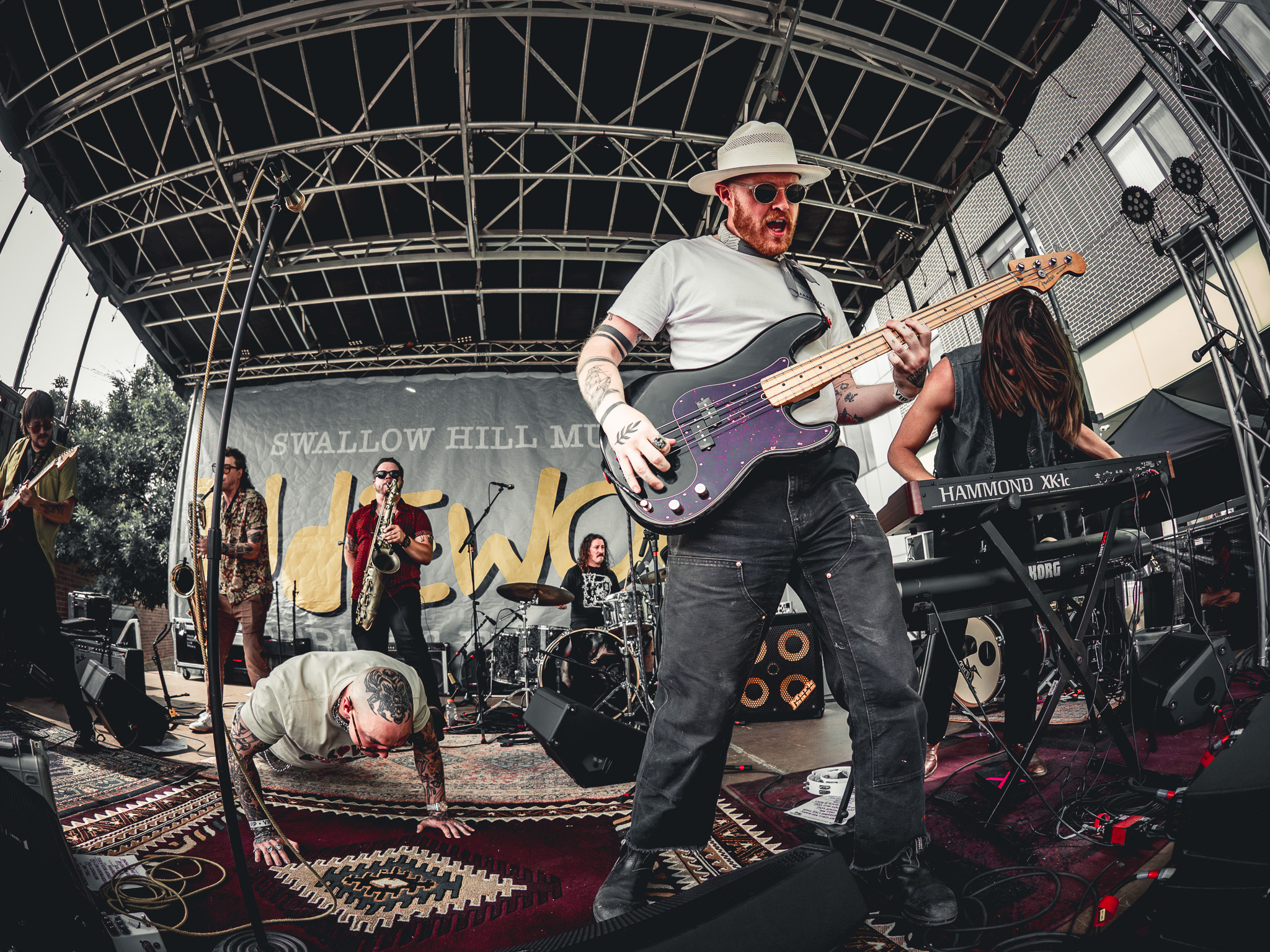Rootbeer Richie headliner for Culture Pop 2026 band playing instruments on stage at a concert. Guitar player with hat is playing while Rootbeer does a pushup on the carpeted stage, with bandmates in back.