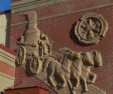 A detailed brick-relief artwork depicting fire station heritage with firefighters, horses, and a fire wagon.