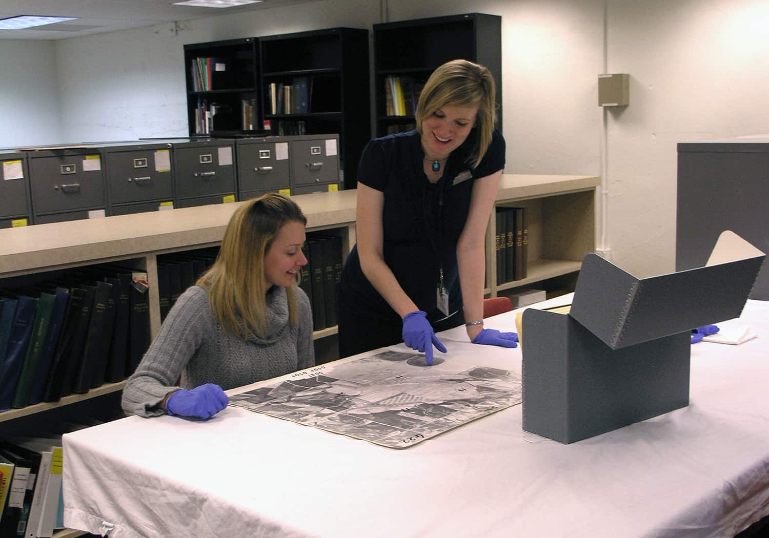 Two individuals engage in examining archival materials in an organized library setting, wearing gloves to handle documents carefully.