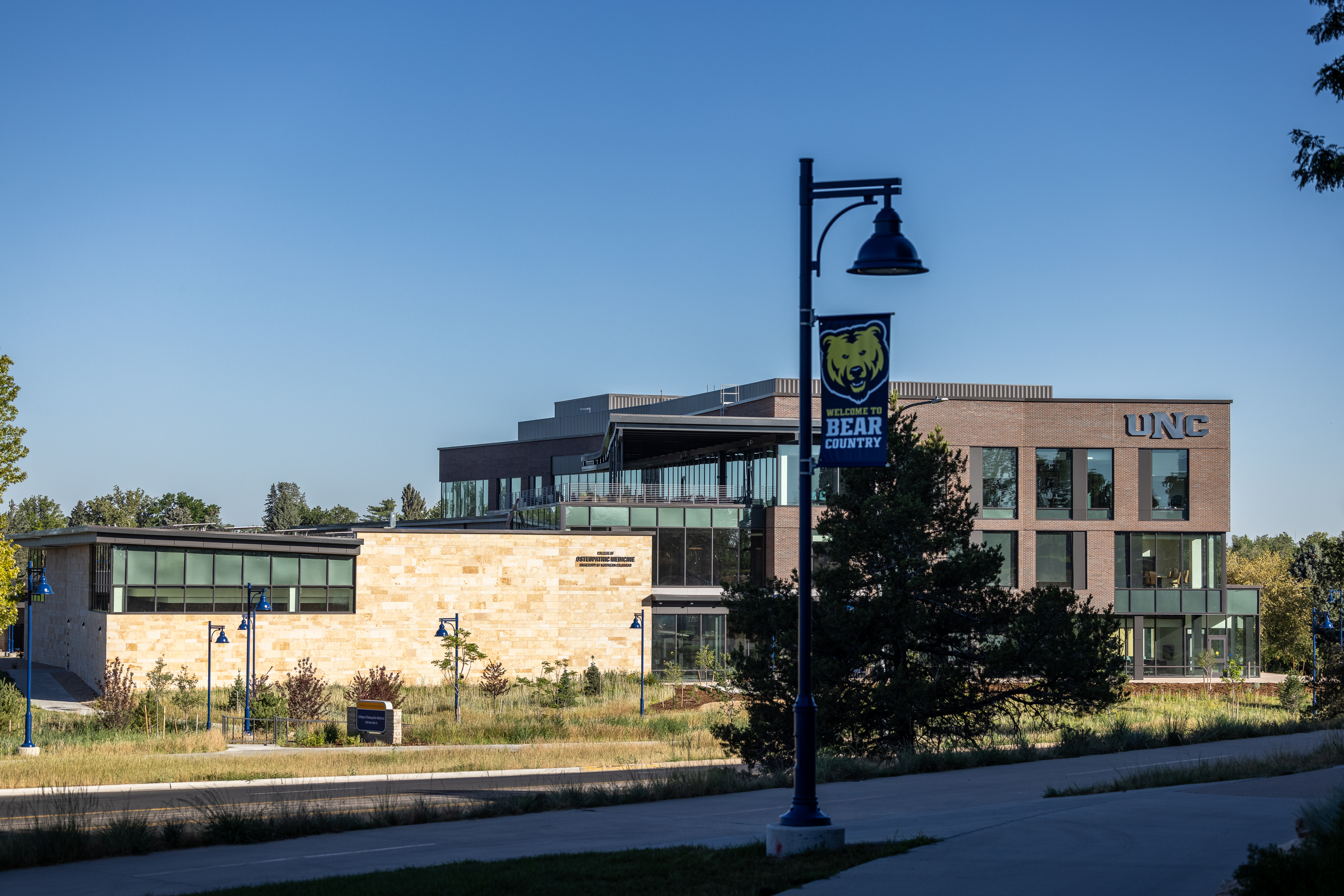 University of Northern Colorado's campus showing the exterior of the College of Osteopathic Medicine in the distance with a lamp post nearby with the UNC Bear on the flag and below saying "Bear Country".