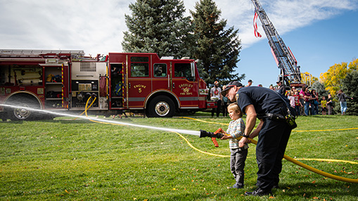 A firefighter helps a young child spray water during a community event featuring a firetruck and demonstrations.