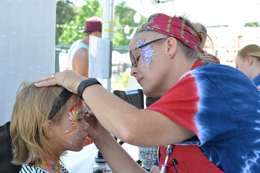 A young girl is enjoying getting her face painted by an artist at the Greeley Arts Picnic, showcasing a lively event atmosphere.