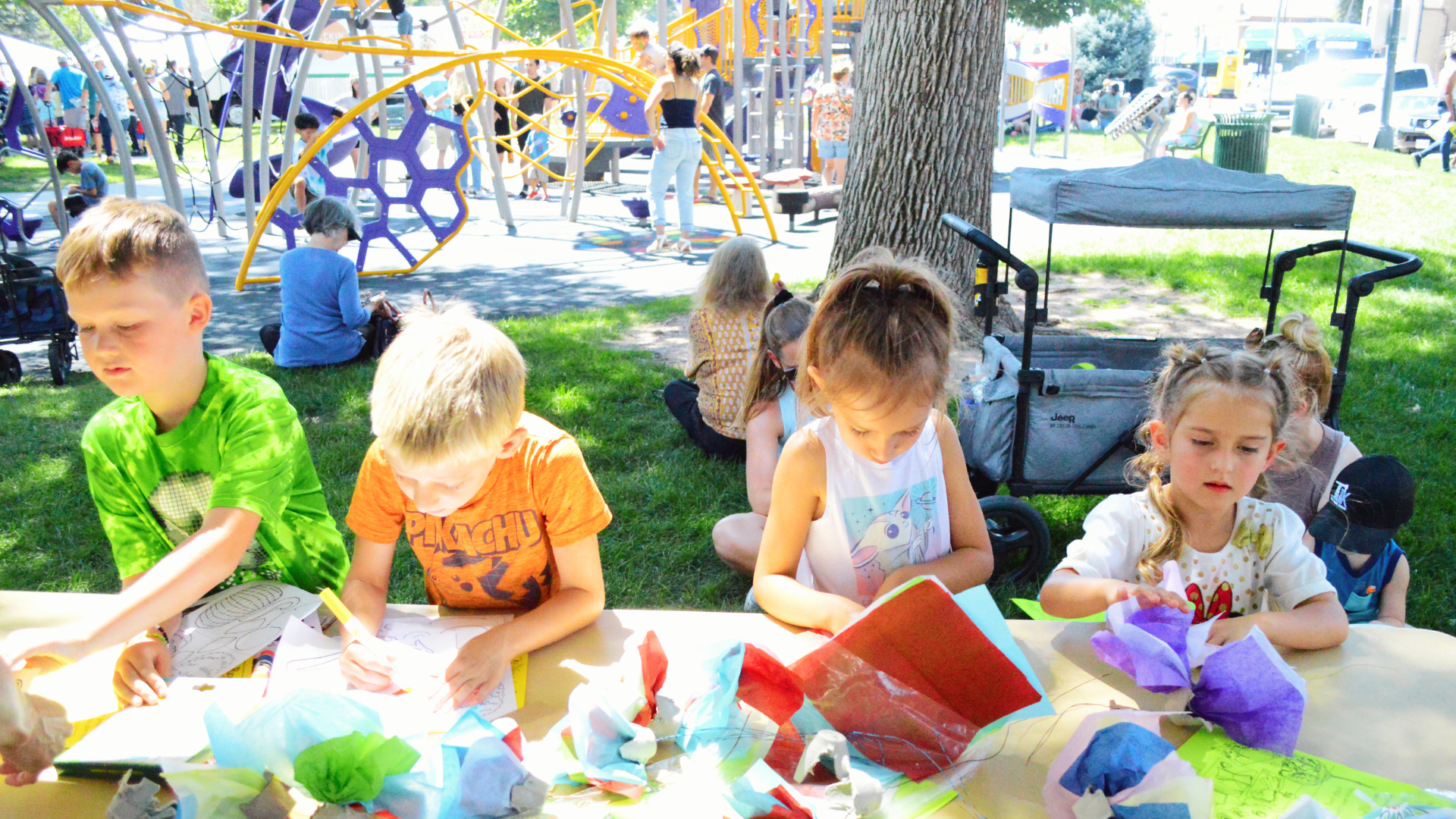 Four kids play with bright art and crafts materials at the Creation Station in Lincoln Park at the Greeley Arts Picnic on a bright sunny day with playground in the distance.