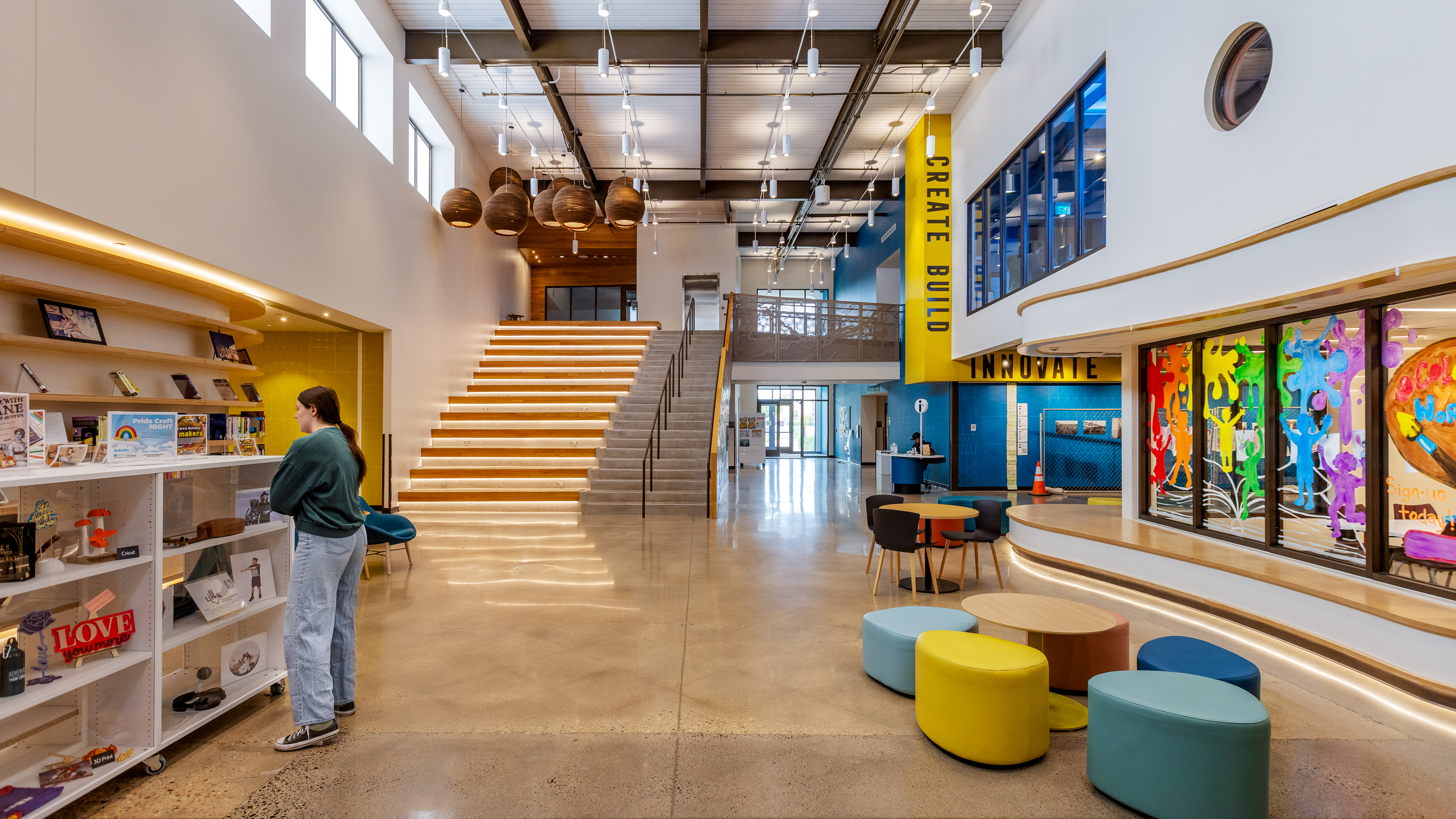 This image depicts the interior lobby of the LINC Library in Greeley, Colorado.  To the right are interior windows adorned with bright red, orange, green, blue and purple stickers in the shapes of children.  In front of those windows are brown tables with light blue, dark blue, and yellow chairs.  A woman is standing to the left of those tables, browsing decorative items on white shelves.  Behind the woman is a staircase taking visitors to the second level of the library.  Modern brown pendant lights hang directly above the staircase.