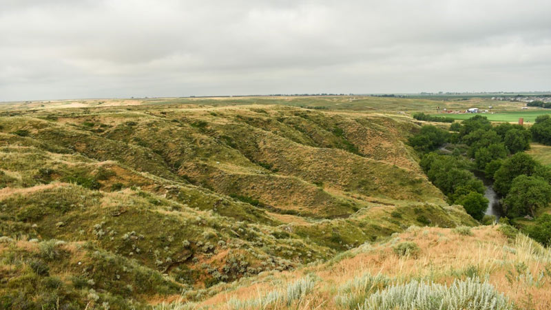 A vast prairie with steep arroyos, a winding river flanked by lush green trees, under a cloudy sky.
