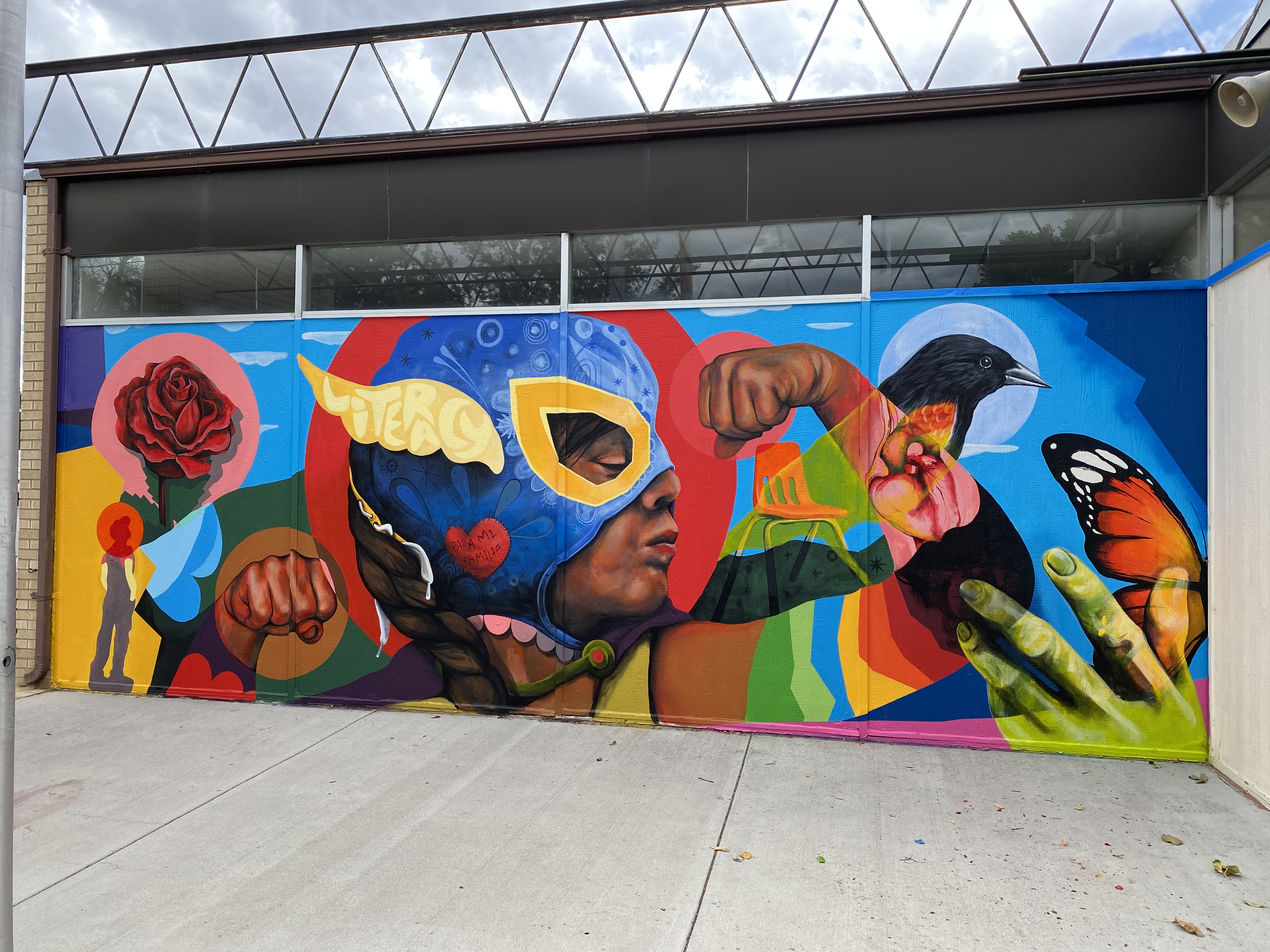 This image depicts a colorful mural painted on the side of a building in downtown Greeley.  The mural shows a young African American child flexing both arms in a show of strength.  The child is wearing a superhero-type mask that is blue with yellow trim and yellow wings bearing the word "literacy".  The child's arm is covered in rainbow stripes.  Surrounding the child are more bold, colorful images that include a Monarch butterfly, a black raven, a red rose, and a hand outstretched to the child.  