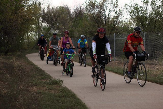 A group of cyclists, including children and adults, enjoy a scenic ride on a paved trail surrounded by nature.