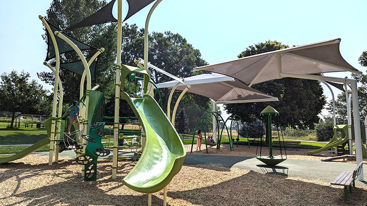 A shaded playground featuring a green slide, wood fiber ground, wood bench, and green trees with children enjoying swings and rope climbing activities.