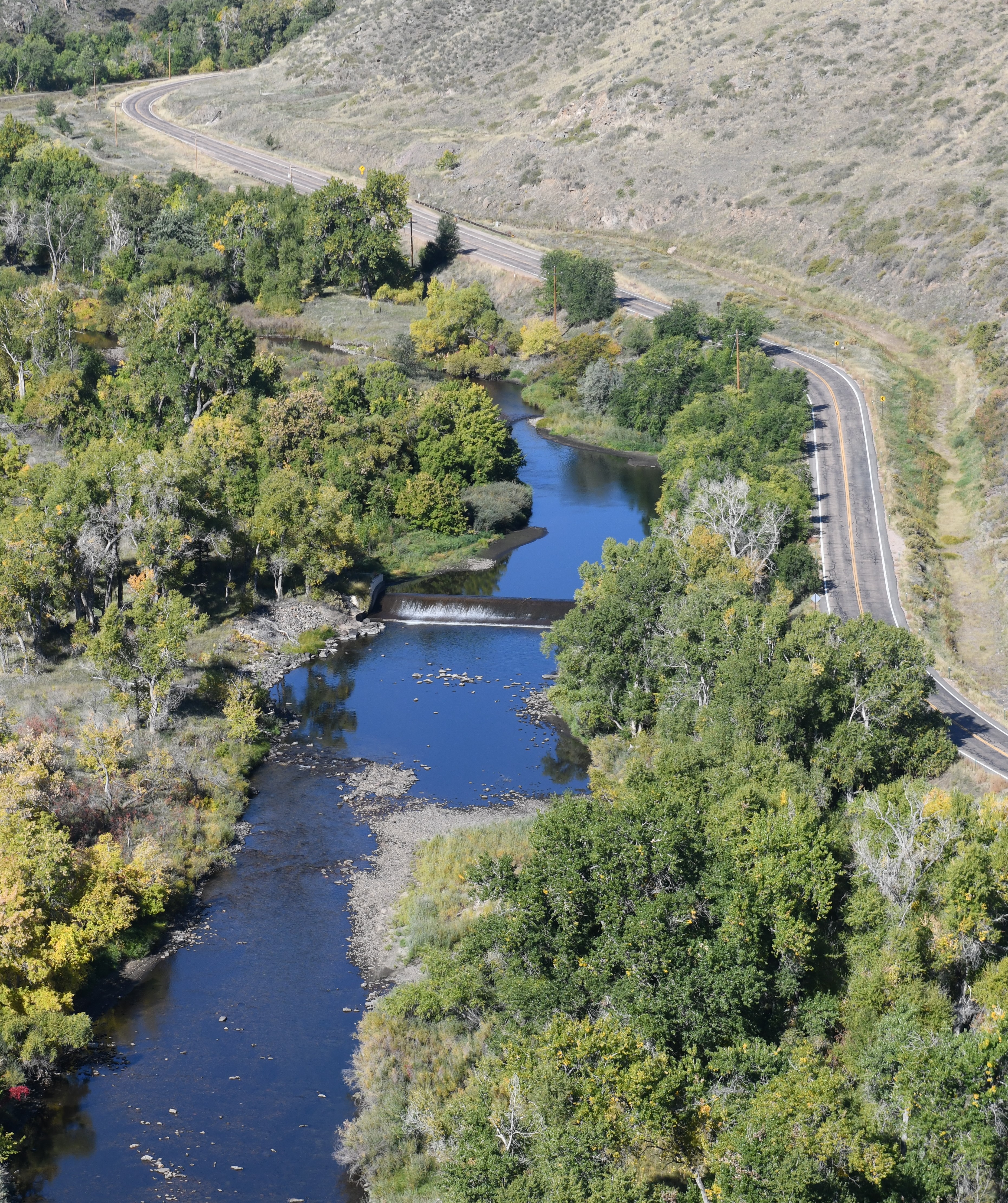 An aerial photograph showing the Cache la Poudre River with a road running alongside it and trees along both banks. There is a diversion structure spanning the width of the river where water flows down a concrete diversion dam, intended to raise the water level of the river upstream to divert water.