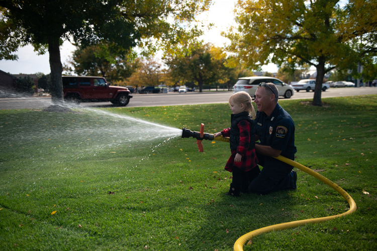 A young child receives guidance from a firefighter on using a firehose during an outdoor community event.