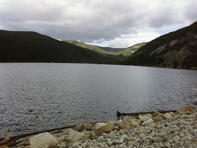 A serene view of Comanche Reservoir surrounded by forested mountains under a cloudy sky.