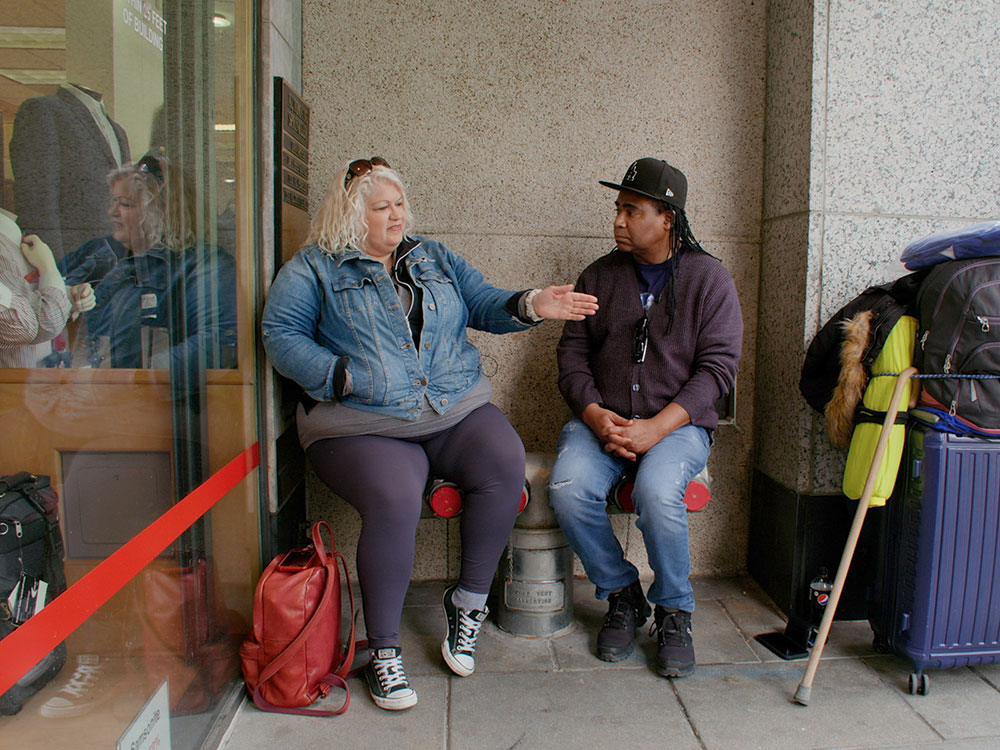 Filmmaker Don Sawyer talks to a woman while seated on chairs on a downtown side walk in front of a large building. They appear to be talking. The woman is pointing towards the street. Luggage and belongings are nearby