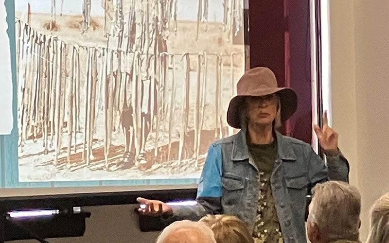 A person dressed as Rattlesnake Kate speaks to a seated crowd at the Greeley History Museum. A black and white photo projected on a screen behind the person shows rattlesnake skins hanging on two lines.
