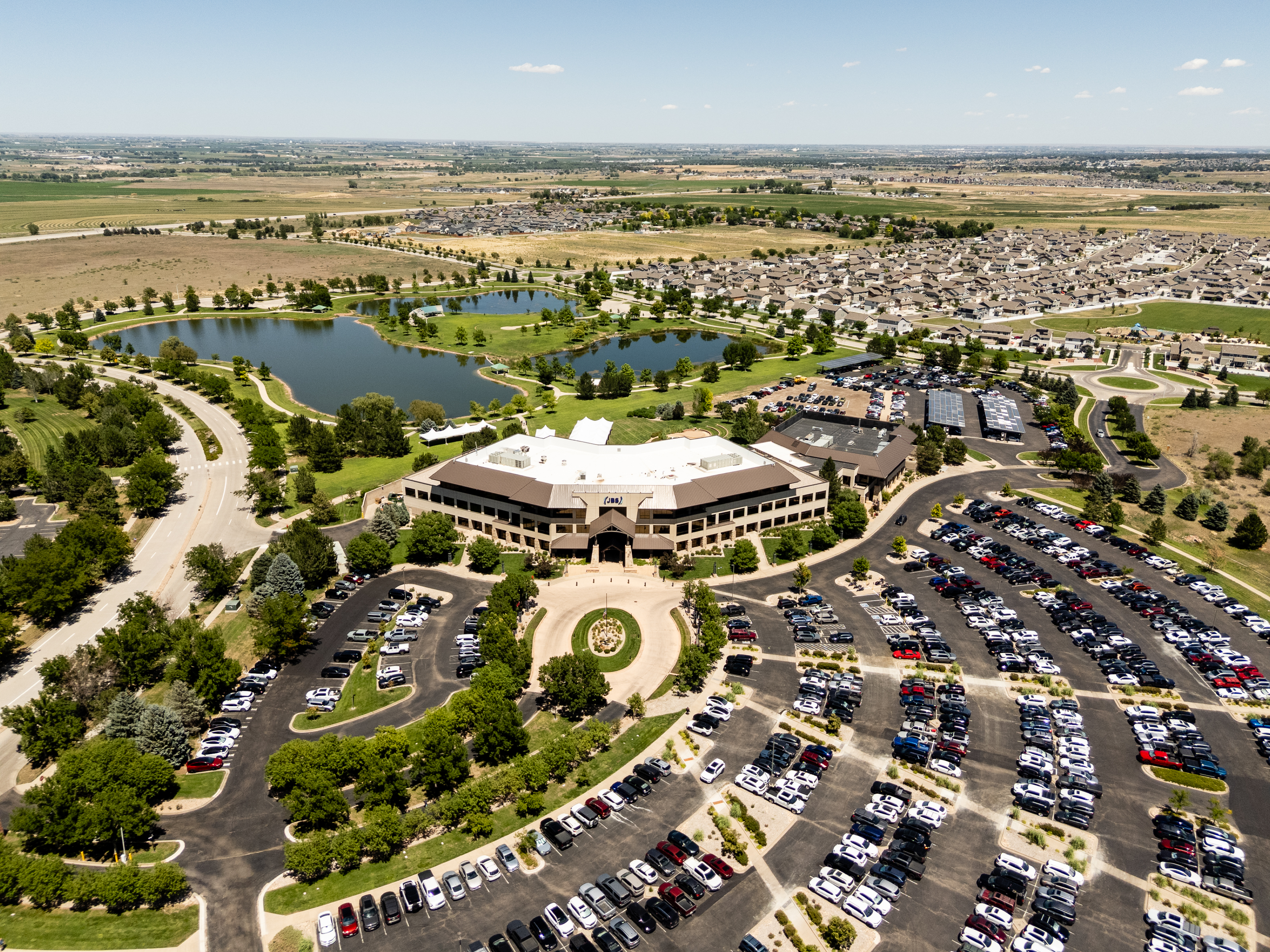 Aerial view of the JBS headquarters building in Greeley, Colorado, with surrounding parking lots, landscaped grounds, nearby ponds, neighborhoods, and open farmland.