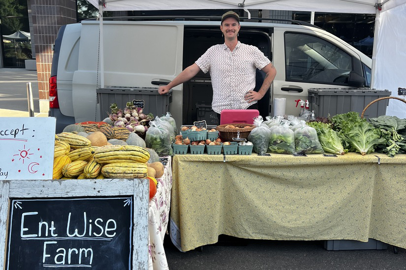 Man with Ent Wise Farm smiling in front of his vegetable spread at the Greeley Farmers' Market. The table has a variety of squash and leafy vegetables. His van is in the background and he is under a white pop-up tent. 