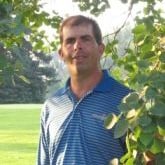 A man wearing a striped blue shirt standing amidst green outdoor scenery with trees and grass in the background.
