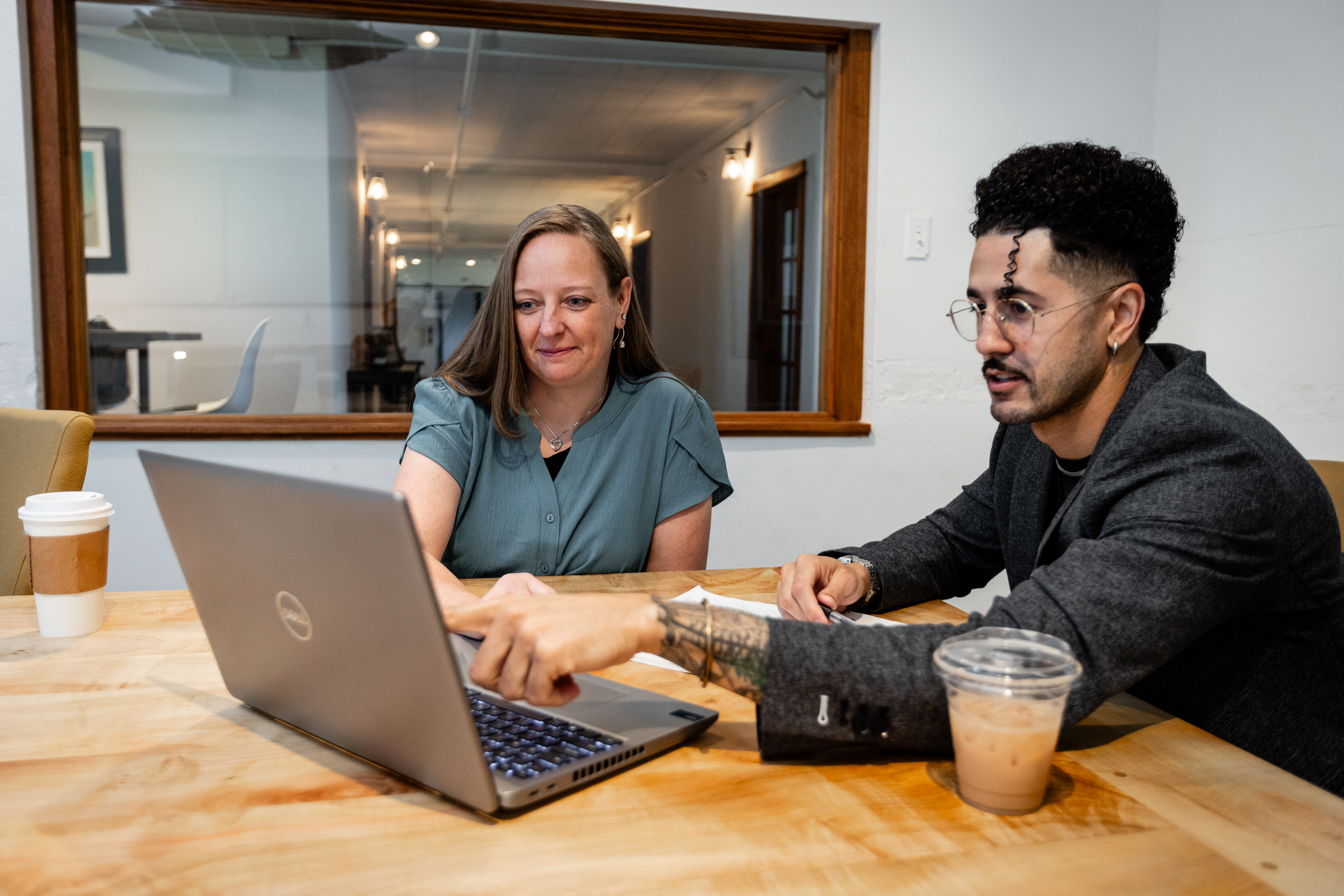 A man wearing glasses and a dark gray sports coat points to his laptop screen while a woman in a green short-sleeve shirt looks on.  The two individuals are sitting at a wooden table and both have coffee cups nearby.  A window behind the woman looks out to additional office space with tables and chairs.