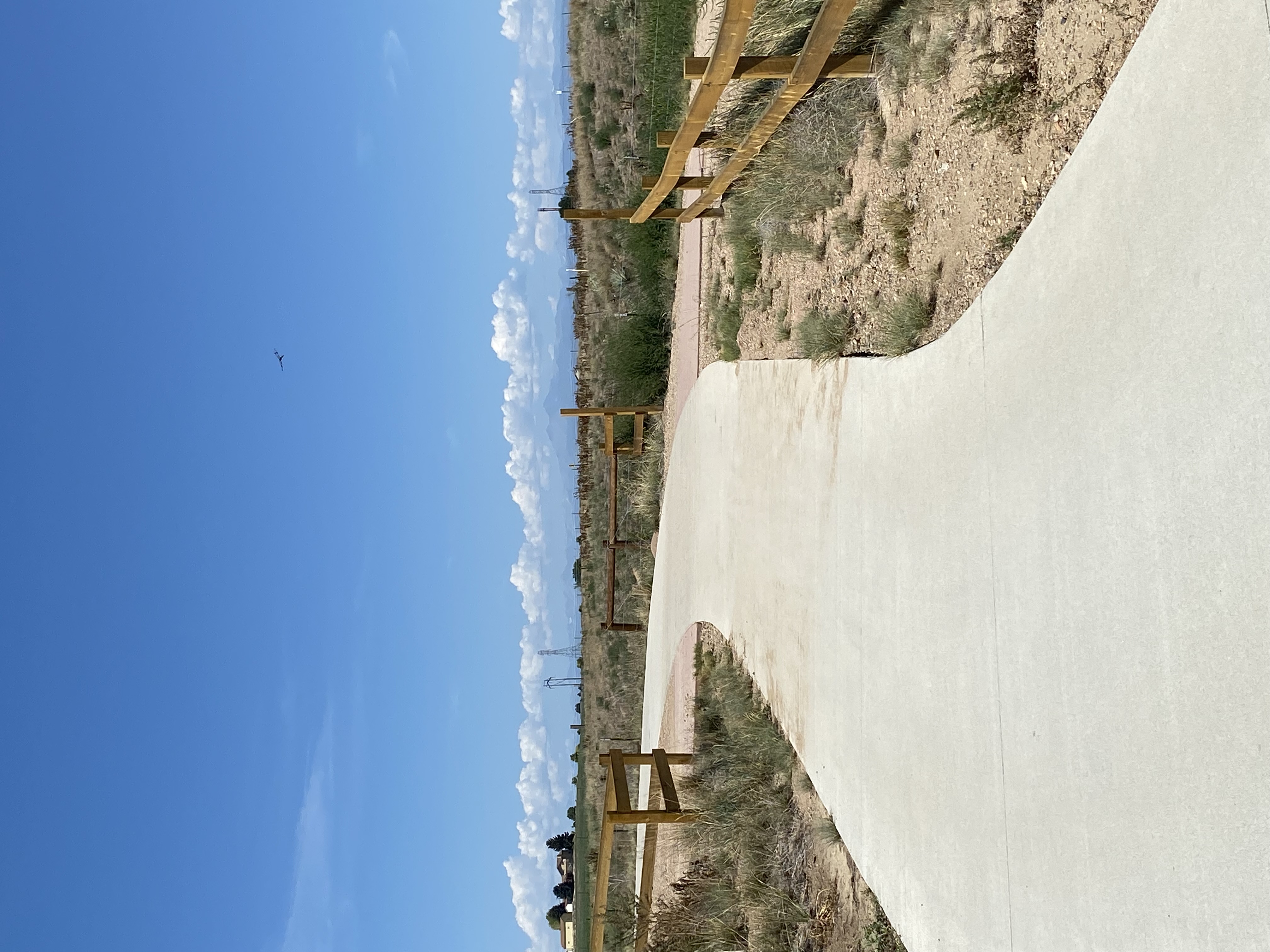 Two concrete trails meet to form a junction that is bordered by wooden fences. In the distance, mountains and clouds are visible. 