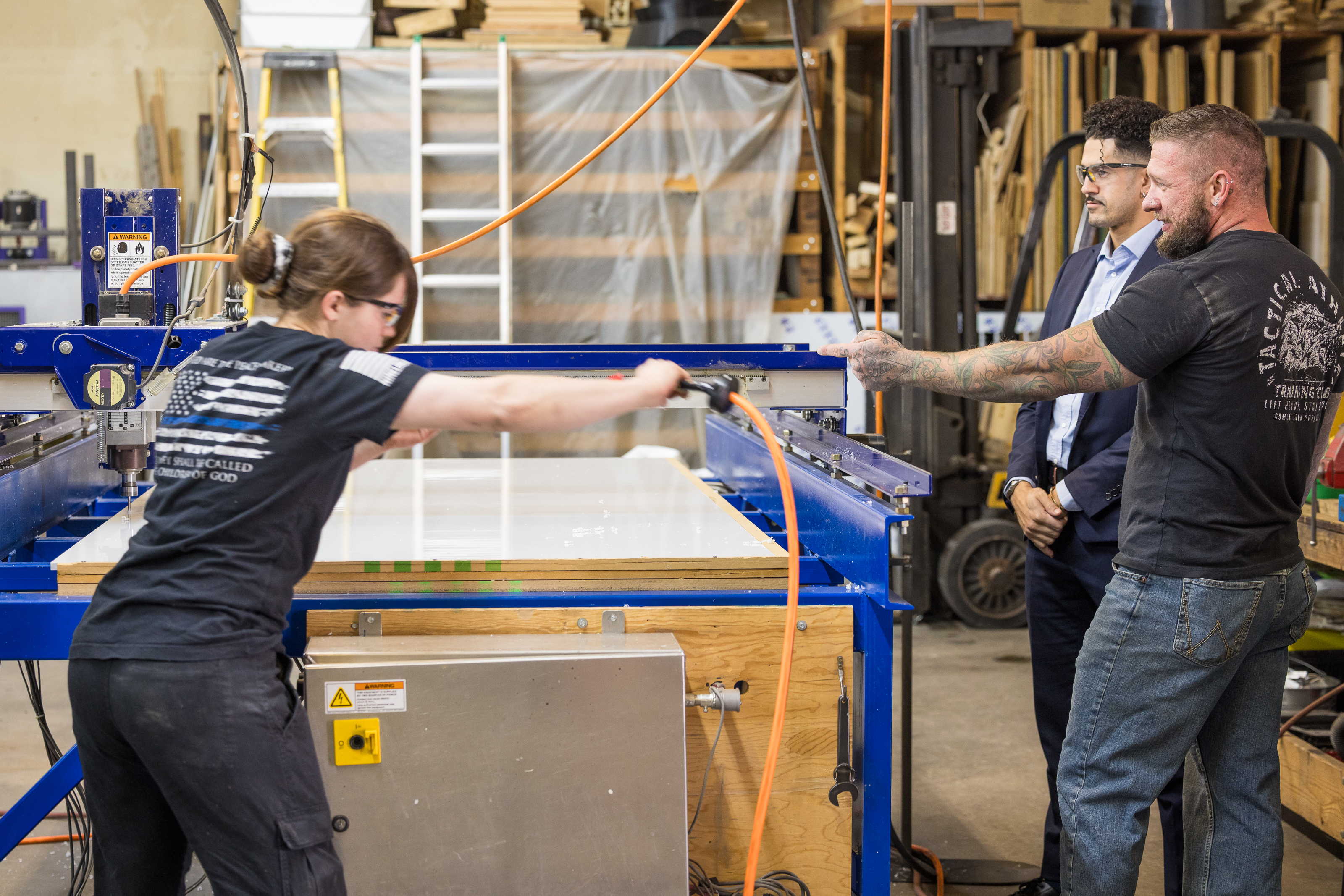 Two workers operate a large industrial machine in a workshop while two men, one in a suit, observe. The setting includes wood materials and equipment in the background, suggesting a manufacturing or fabrication environment.