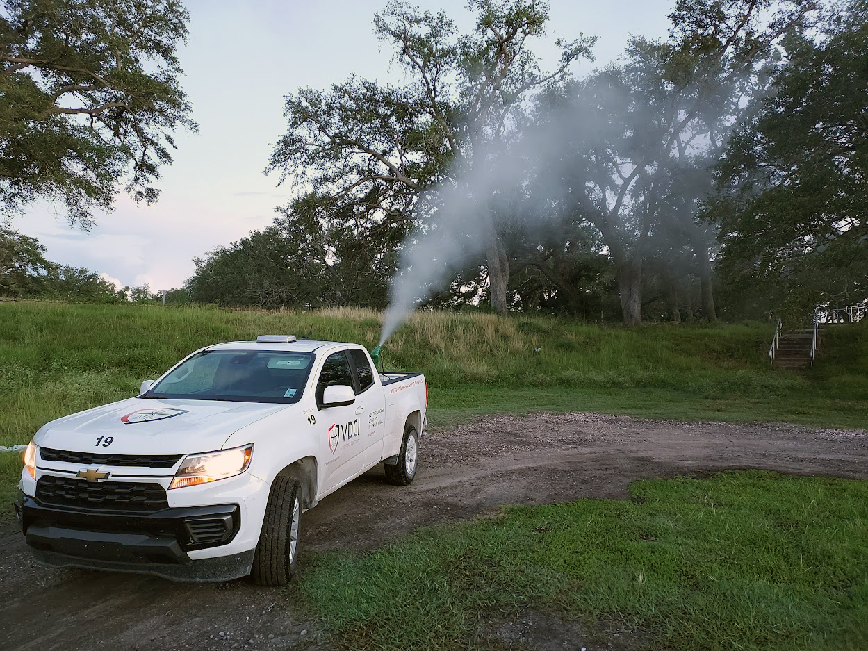A VDCI company truck sprays an insecticide mist behind it as it drives along a dirt road in a wooded area
