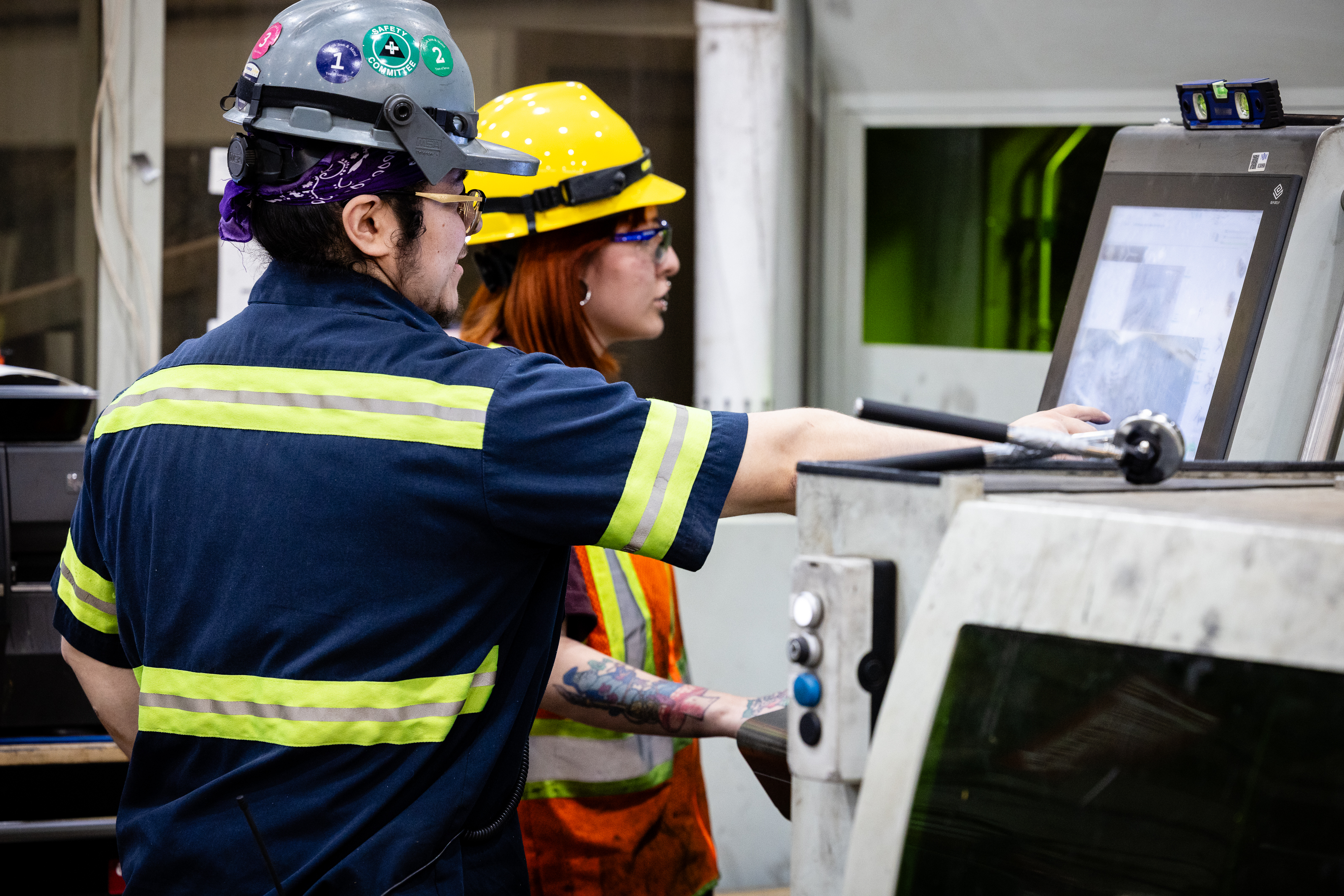 Two workers wearing safety helmets, protective glasses, and reflective gear operate a large industrial machine, focusing on a control screen inside a manufacturing facility.