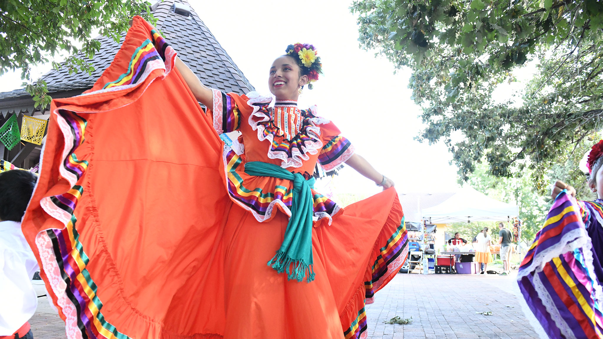 Latina with flowers in her hair holding out the sides of a traditional Mexican dress like a fan during an outdoor daytime event