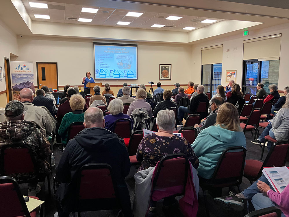 A person stands behind a podium giving a presentation in front of a room full of people seated in chairs. There is a screen on the wall with a Powerpoint presentation shown.