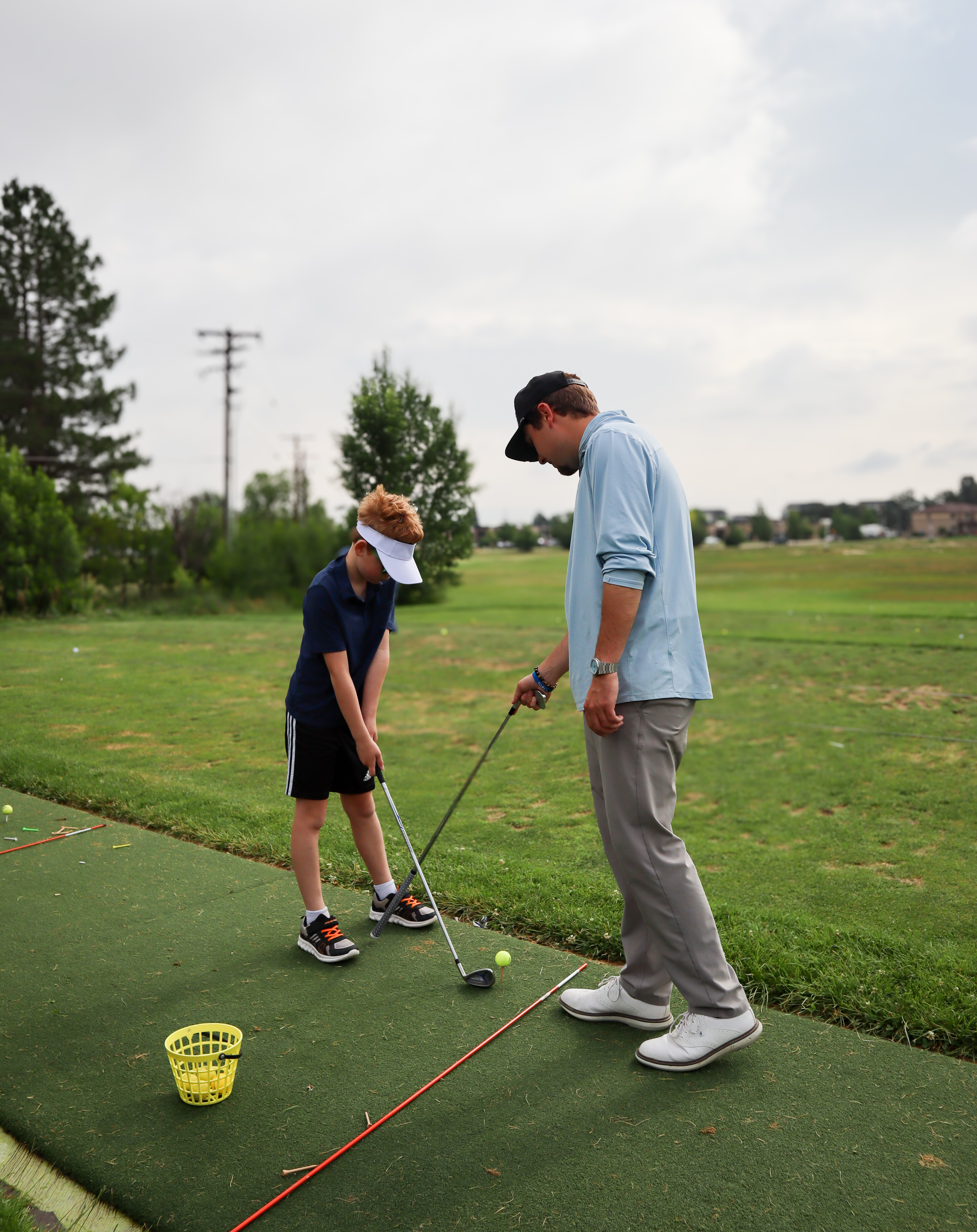 Golf instructor teaching a child proper set up technique on a turf mat at an outdoor driving range. 