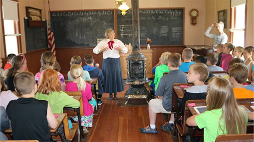 A class of young students attentively listening to a teacher demonstrating in a historic classroom setting, featuring chalkboards, a stove, and traditional furnishings.