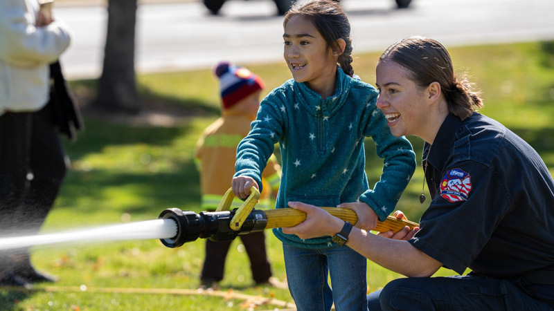 A female firefighter helps a young girl use a fire hose during a community event while another child dressed in a firefighter outfit participates in the background.