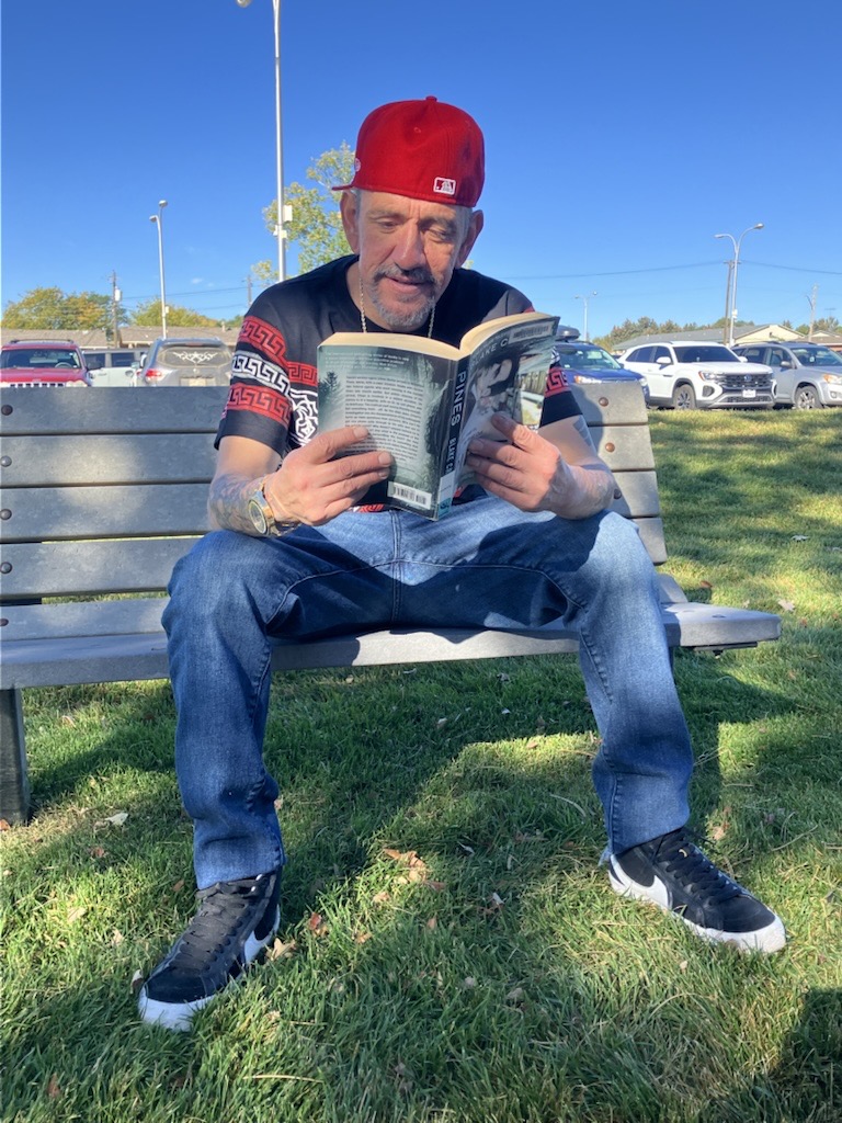 Travis sitting in a park on a bench holding a book open and reading it. 