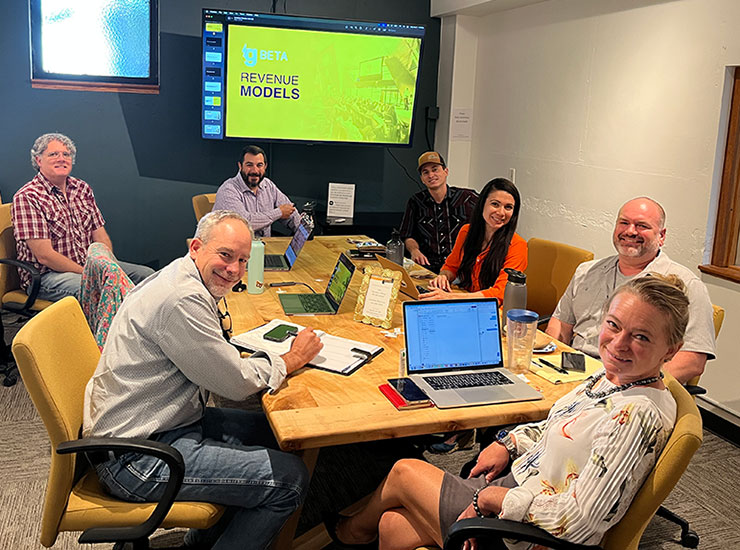 gBETA Greeley cohort participants and mentors smile for a photo around a conference table during a Revenue Models session. A presentation slide is visible on the screen at the back of the room.