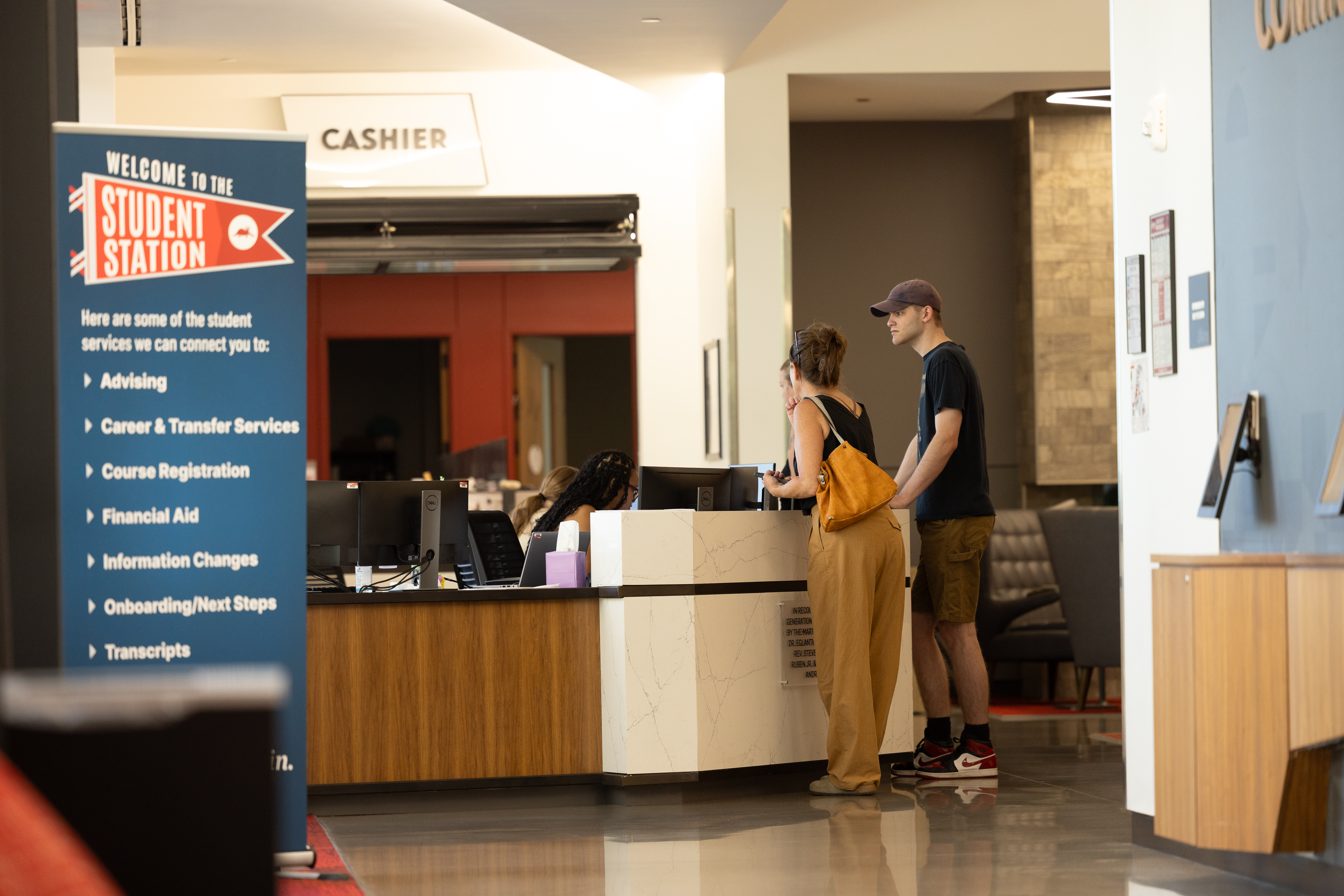 Students speak with staff at the Aims Community College Student Station service counter, with signage for advising, financial aid, registration, transcripts, and other student services.