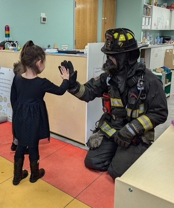 Greeley Firefighter kneeling in black and yellow bunker gear, helmet, and breathing apparatus, giving a high five to a little girl in black clothing on a red, orange, and yellow carpet with two children watching inside a classroom with green walls and two wooden doors in the background. 