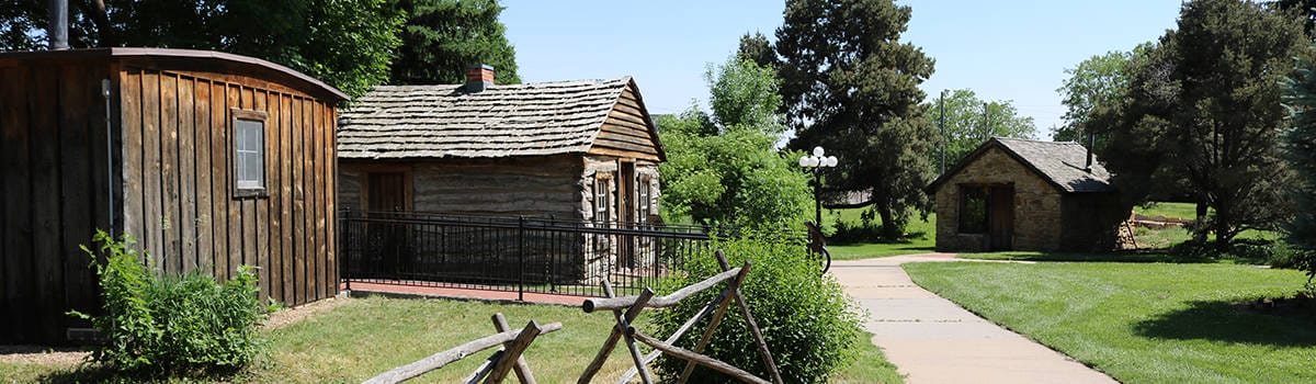 A serene view of Centennial Village showcasing rustic log cabins surrounded by lush greenery and pathways.