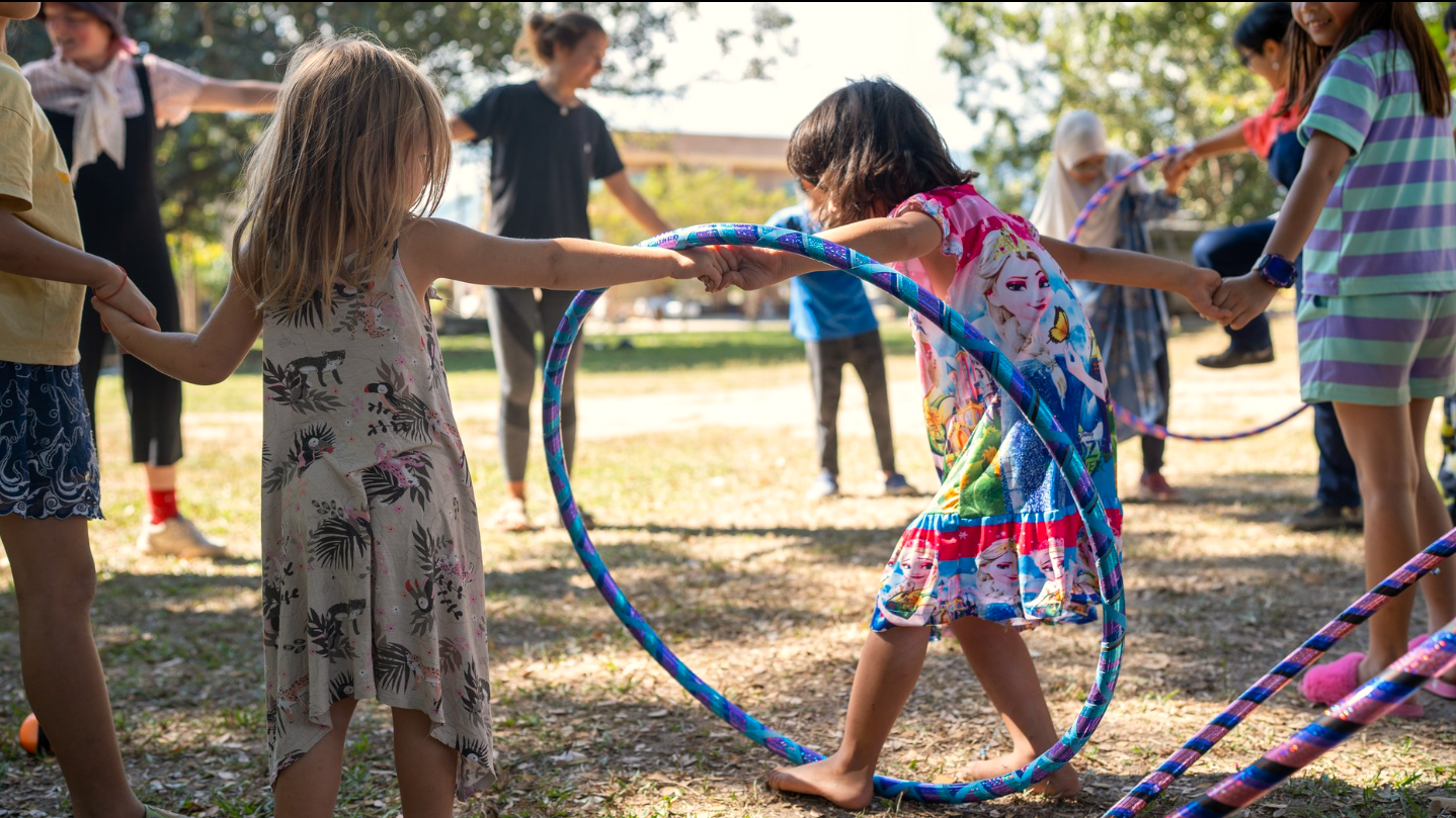 Kids holding hands in a circle playing with hula hoops during a Spoon Circus workshop outside in a park.