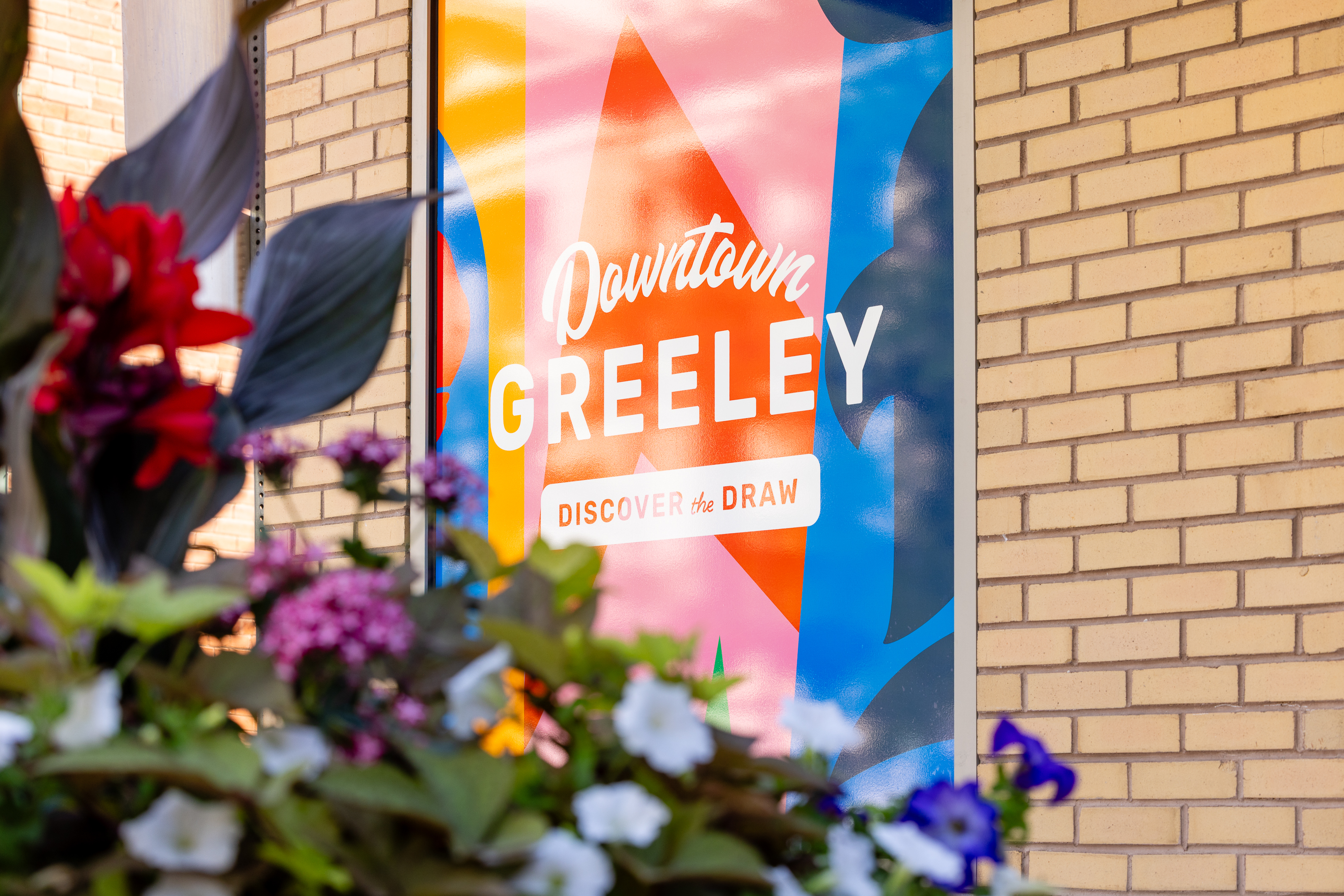 Colorful “Downtown Greeley” sign on a brick wall with flowers in the foreground.