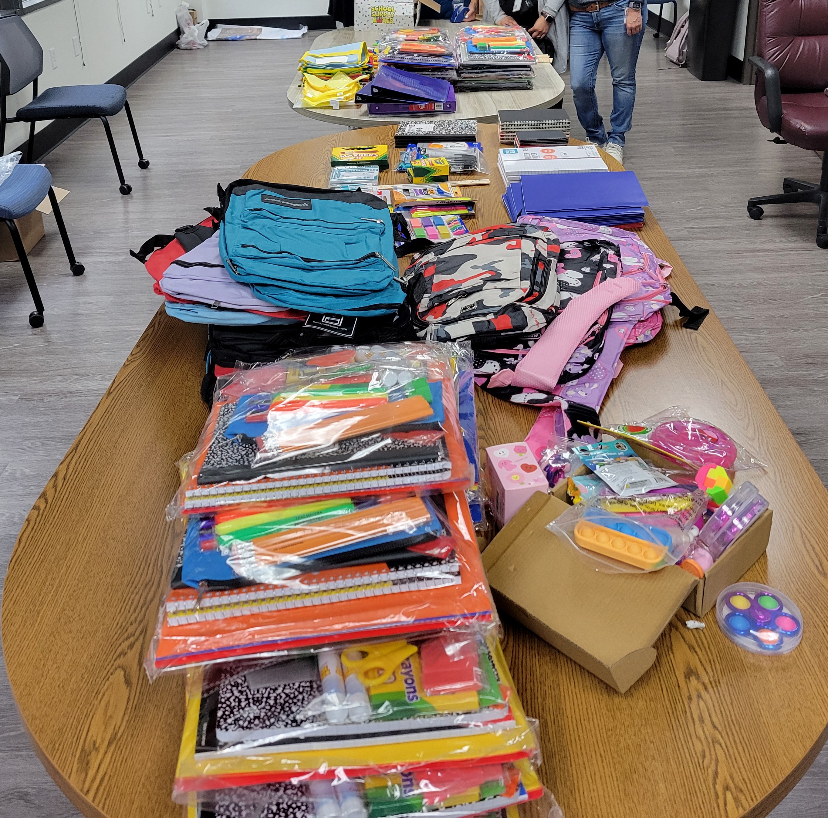 Two long wooden tables in an office with school supplies such as notebooks, highlighters, backpacks, fidget toys, markers, colored pencils, folders, rulers and binders. 