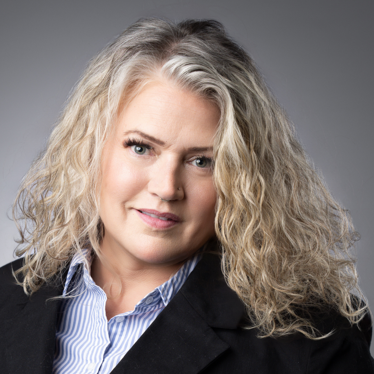 This image depicts a professional headshot of Real Estate Manager Kirsten Muncy.  Kirsten is shown in a dark suit jacket and a blue and white vertical striped dress shirt.