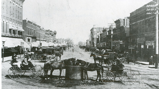 A historical view of downtown streets featuring horse-drawn carriages, showcasing a bustling early local business environment.