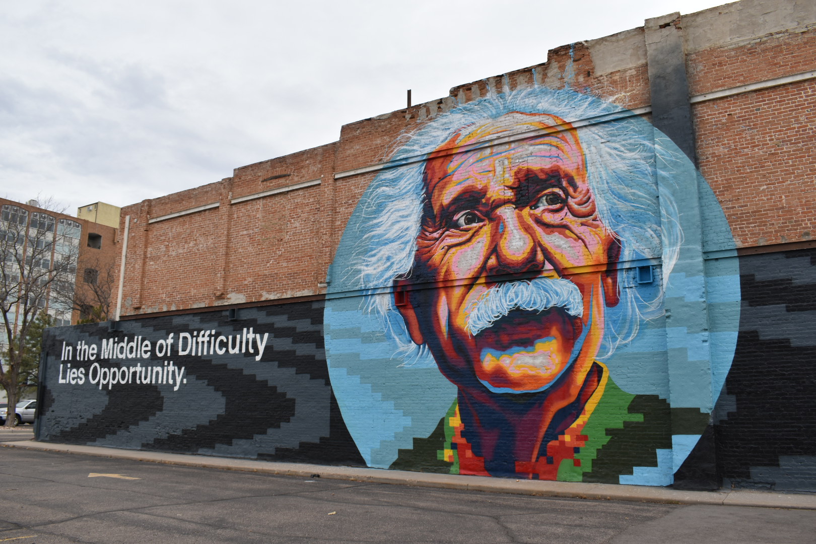 This image depicts a mural painted on the side of a red brick building in downtown Greeley.  The mural depicts an artistic rendering of the face of Albert Eistein, glancing into the distance, inside of a blue circle.  A gray banner is behind the blue circle and contains the words "In the Middle of Difficulty Lies Opportunity."