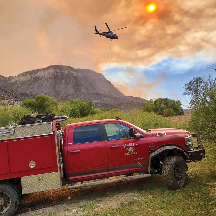 Red Greeley Fire Department Brush Truck with Tool box outdoors on a grassy slope in front of a mountainous area with a helicopter flying above in a smokey sky. 