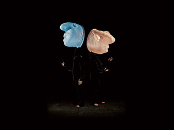 This image showcases two performers, each wearing dramatic headpieces in contrasting colors, set against a dark background, expressing creative visual artistry.