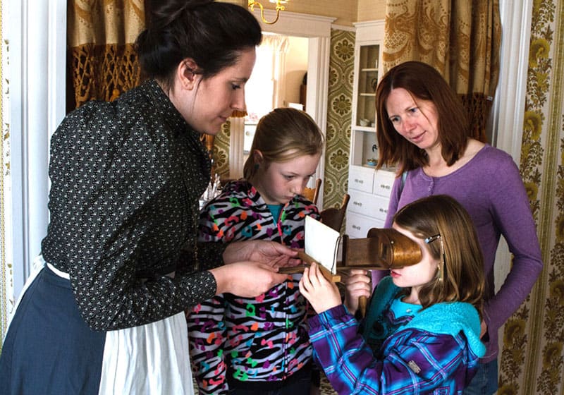 An educator demonstrates historical tools to children in a recreated period room setting at Centennial Village.