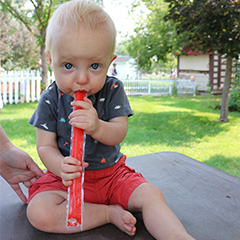 A young child sits on a wooden table holding a popsicle in a lush green yard with a white picket fence and trees in the background.